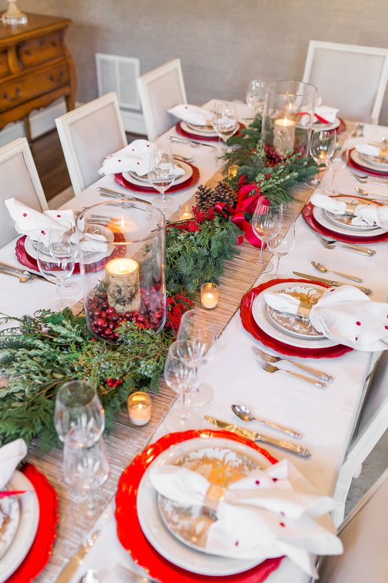 إعداد طاولة عيد الميلاد المشرقة والأنيقة مع الخضرة والتوت الأحمر والشموع المغلفة باللحاء والشواحن الحمراء والمناديل المنقطة