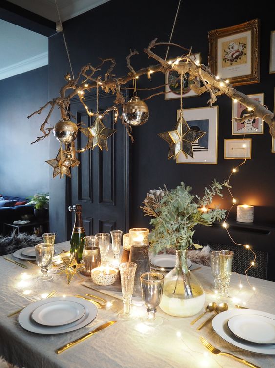 طاولة عيد الميلاد الأنيقة مع فرع مع الحلي والنجوم المعلقة فوق الطاولة والأضواء والمساحات الخضراء على الطاولة وأدوات المائدة الذهبية