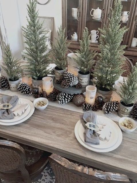 منظر طاولة أنيق لعيد الميلاد مع أكواز الصنوبر المعتادة والثلجية ، وأشجار عيد الميلاد المحفوظة بوعاء ، والبياضات المحايدة وأشجار عيد الميلاد في إعدادات المكان