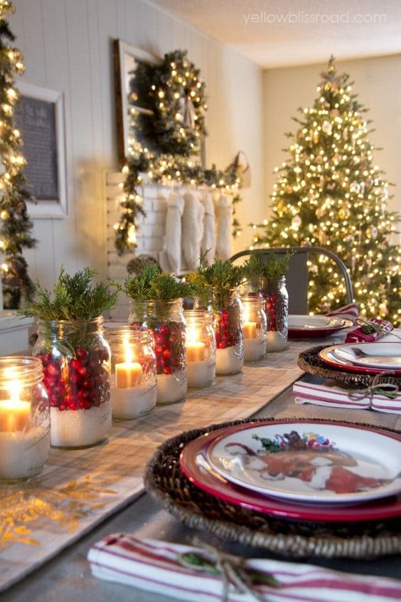طاولة عيد الميلاد المريحة مع المفارش المنسوجة والمرطبانات ذات الثلج الزائف والتوت والمساحات الخضراء والشموع والألواح المطبوعة مشرقة ورائعة