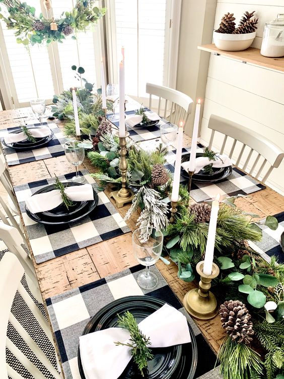 طاولة عيد الميلاد في مزرعة مع عداء دائم الخضرة وصنوبر ، وشموع طويلة ورفيعة ، ومفارش مربعة من الجاموس ، وألواح سوداء