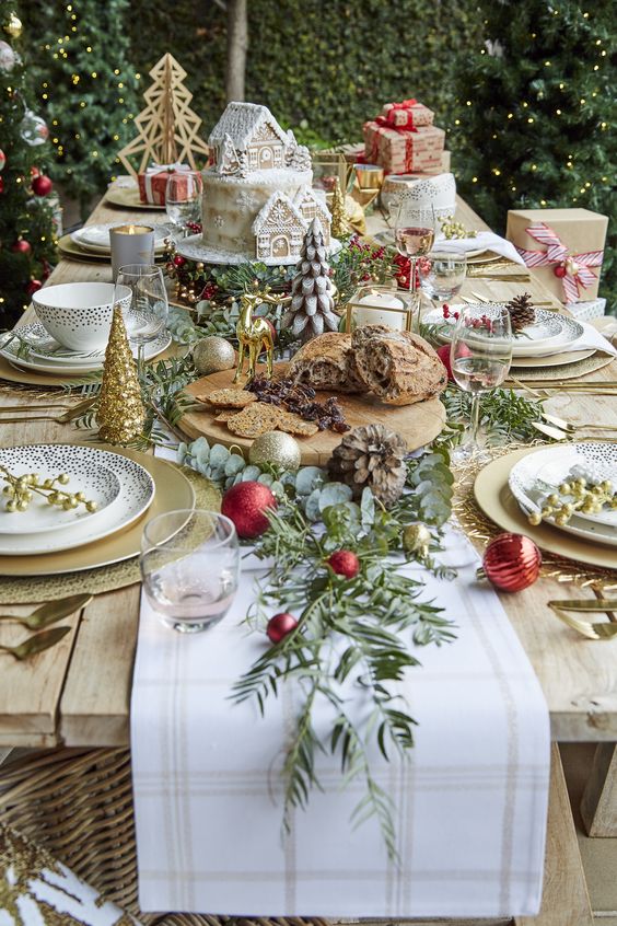 طاولة عيد الميلاد الرائعة مع المفارش والشواحن الذهبية ، وأشجار عيد الميلاد الصغيرة والزخارف ، والمساحات الخضراء ، والزخارف الحمراء ، وكعكة عيد الميلاد الرائعة