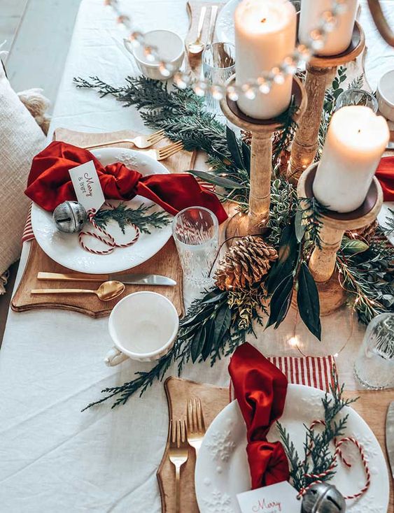 طاولة عيد الميلاد الجميلة مع المساحات الخضراء وأكواز الصنوبر وحوامل الشموع الخشبية مع شموع عمودية ومناديل حمراء وألواح خشبية مقطوعة وأجراس