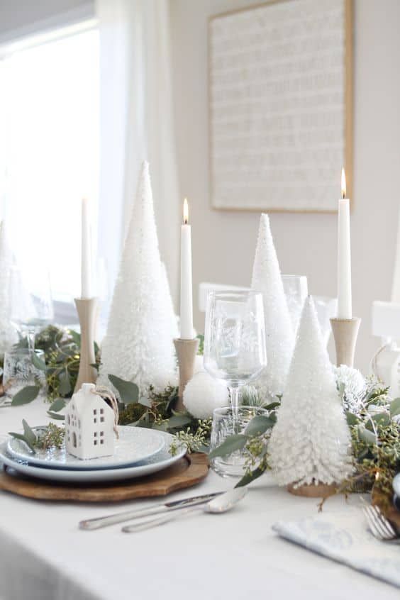 طاولة عيد الميلاد الجميلة مع أشجار الفرشاة البيضاء ، والشموع البيضاء في حاملات الشموع الخشبية ، والمفارش الخشبية ، والمنازل البيضاء والأوكالبتوس الطازج