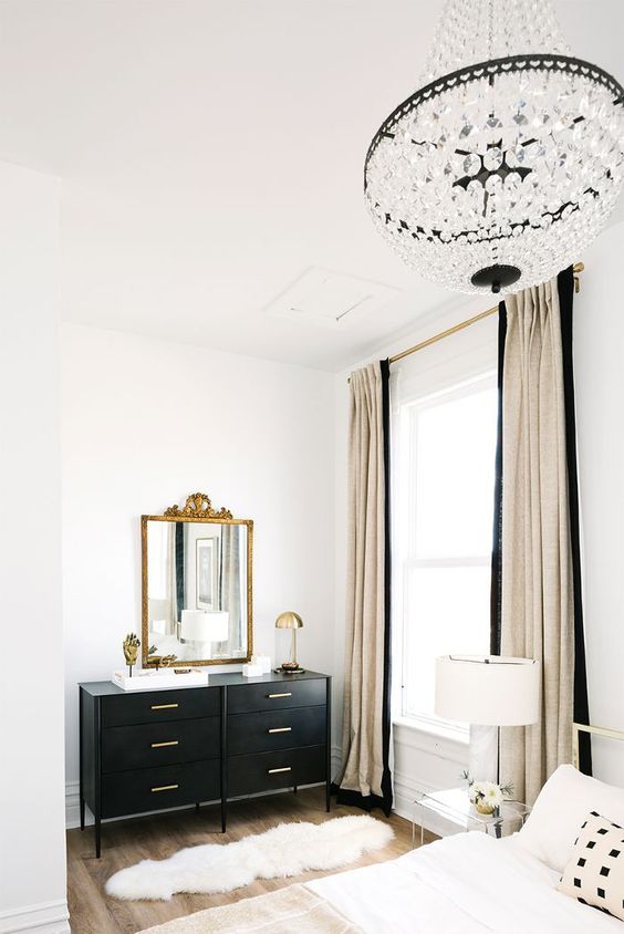 a glam Parisian bedroom in neutrals and black, with a crystal chandelier, a black sideboard, a gold mirror and lamps