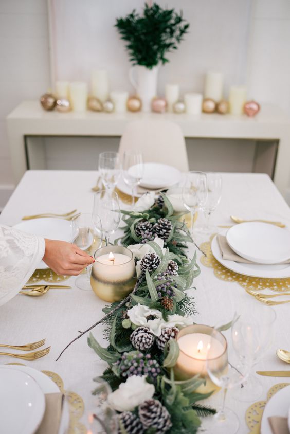 طاولة شتوية ساحرة مع المفارش الذهبية ، دائمة الخضرة ، أزهار بيضاء وعداء كوز الصنوبر الثلجي ، شموع وأدوات مائدة ذهبية