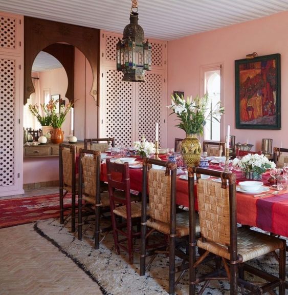 مساحة مغربية جريئة لتناول الطعام بجدران وردية وطاولة طويلة مع مفرش طاولة أحمر وكراسي منسوجة ومصباح معلق جريء