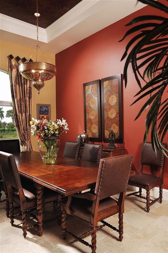 مساحة مظلمة لتناول الطعام بجدار أحمر اللون ، وطاولة منحوتة ثقيلة داكنة ، وكراسي جلدية ومصباح مغربي معدني يضفي أجواءً مزاجية.