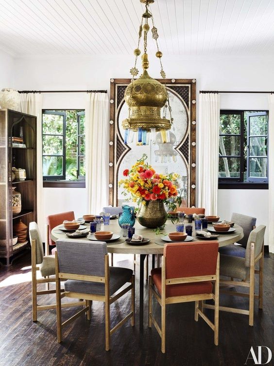 غرفة طعام جميلة مستوحاة من الطراز المغربي مع طاولة مستديرة وكراسي ملونة هادئة ومصباح معلق معدني جريء ومرآة كبيرة في إطار خشبي منحوت