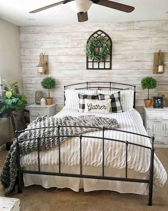 A Farmhouse Bedroom With A Whitewashed Wood Accent Wall A Black Metal Bed Whitewashed Nightstands Potted Plants And A Greenery Wreath 