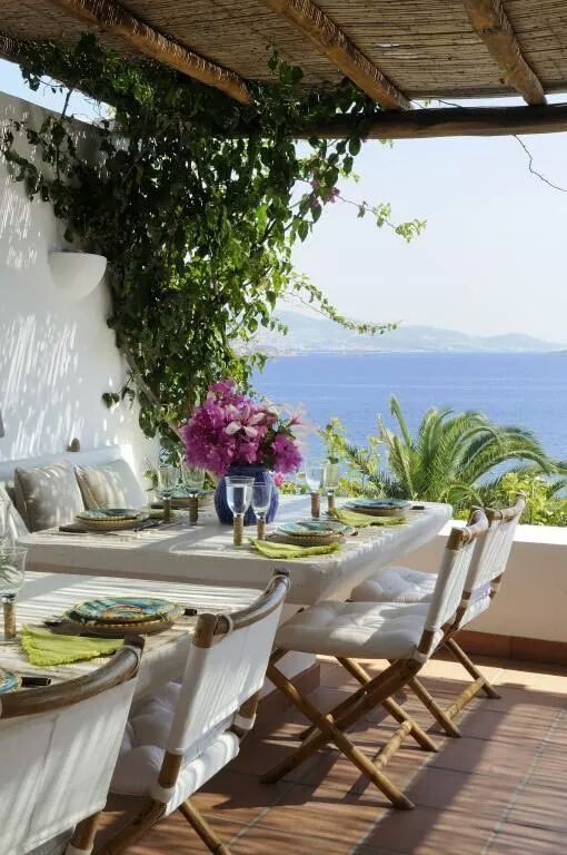 مساحة لتناول الطعام على طراز البحر الأبيض المتوسط ​​مع إطلالة ، مع أريكة مدمجة وطاولات بيضاء وكراسي قابلة للطي مع تنجيد أبيض وأزهار ومساحات خضراء