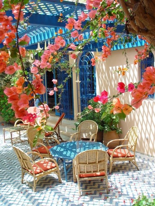 مساحة مشرقة لتناول الطعام في البحر الأبيض المتوسط ​​مع طاولة مستديرة زرقاء وكراسي من الخيزران مع وسائد مرجانية وأزهار زاهية فوق المساحة وإطارات نوافذ زرقاء