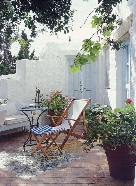 زاوية خارجية صغيرة من البحر المتوسط ​​مع أريكة مع وسائد ووسائد بيضاء ، وكراسي قابلة للطي ، وطاولة معدنية ، وأزهار زاهية في المزارعون