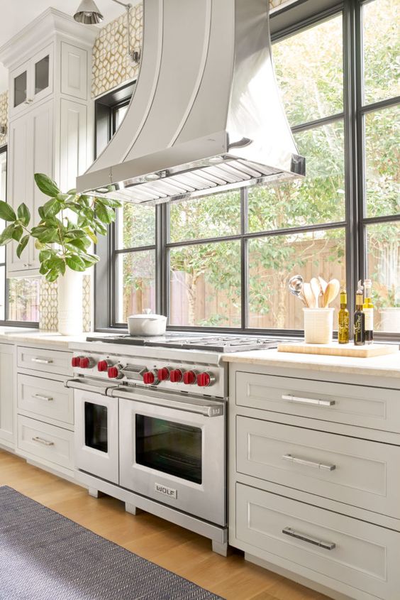 a white vintage farmhouse kitchen with shaker style cabinets, a large metal hood and a big window that opens on the garden