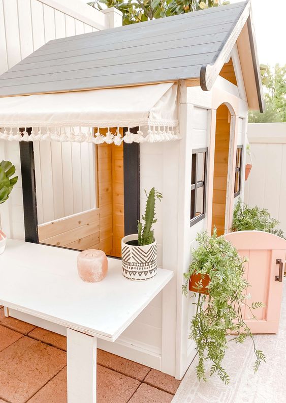 منزل جميل للأطفال باللونين الأبيض والأسود مع باب نصف قرنفلي ، مع أصص خضراء ونباتات ، مع طاولة كبيرة بجانبه