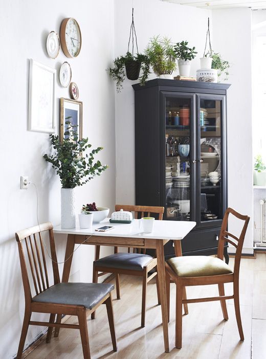 a stylish modern dining nook with a table, blue and green chairs, greenery in pots and vases and a gallery wall with clocks and plates