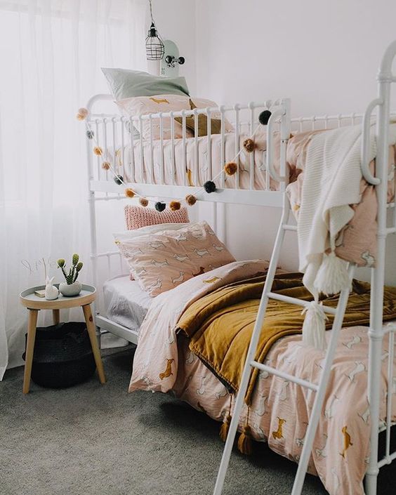 غرفة نوم مشتركة بوهو للفتيات مع سرير بطابقين معدني ، وأغطية فراش باللون الوردي والخردل ، وطاولة جانبية مستديرة ومصابيح معلقة