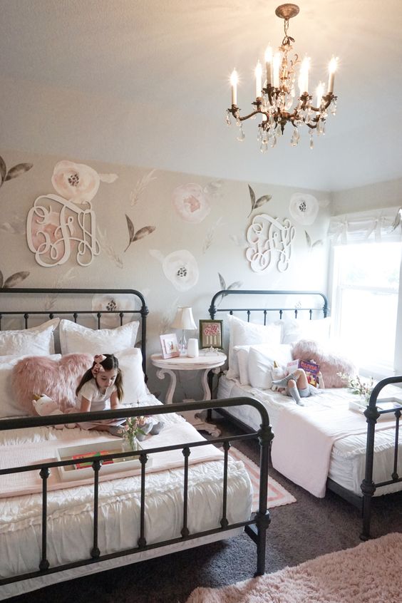 منزل مزرعة أنيق مشترك بغرفة نوم للفتيات مع جدار بلمسات أزهار وحروف أحادية ، مع أسرّة معدنية وأسرّة بيضاء وزهرية ، وطاولة مستديرة كمنضدة