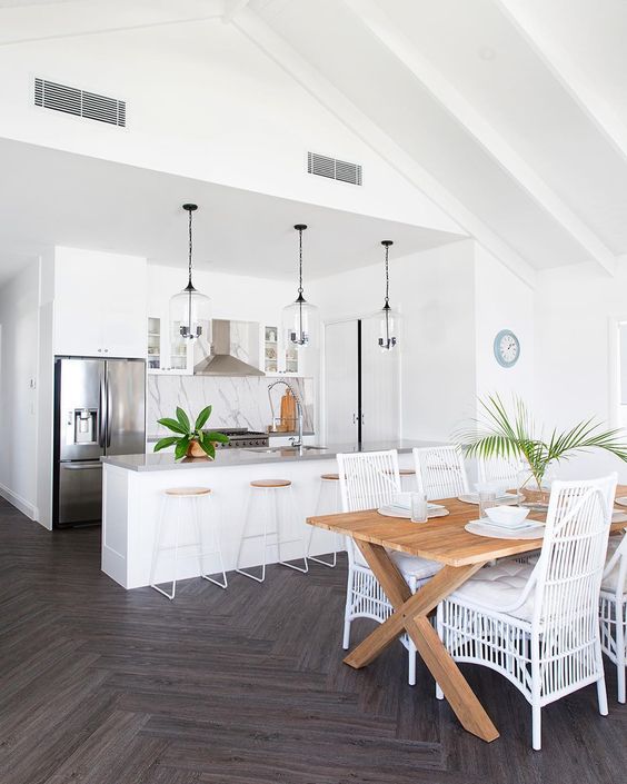 A White Tropical Kitchen With A Marble Backsplash Glass Pendant Lamps Wood And Metal Stools And A Dining Space With A Wooden Table And White Rattan Chairs 