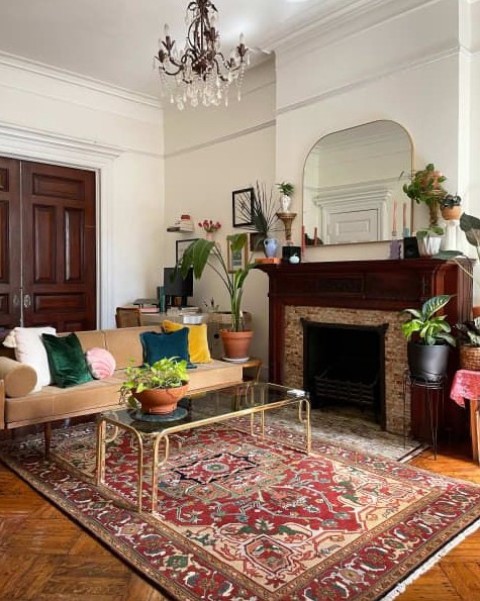 a refined eclectic living room with a fireplace, a beige sofa, bright pillows, a bold rug and a glass coffee table, a crystal chandelier