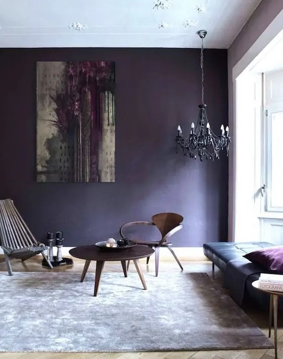 an eclectic living room with a black leather daybed, a couple of wooden chairs and a table, a black chandelier and a deep violet accent wall