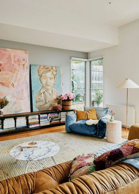 an eclectic living room with a blue and mustard chair, a beige sofa, colorful pillows, a bookshelf, a marble table and artwork