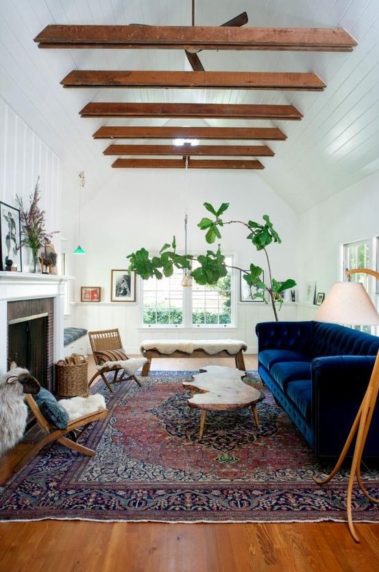 an eclectic living room with a navy sofa, a fireplace, wooden beams on the ceiling, a living edge table, a bench and chairs with faux fur