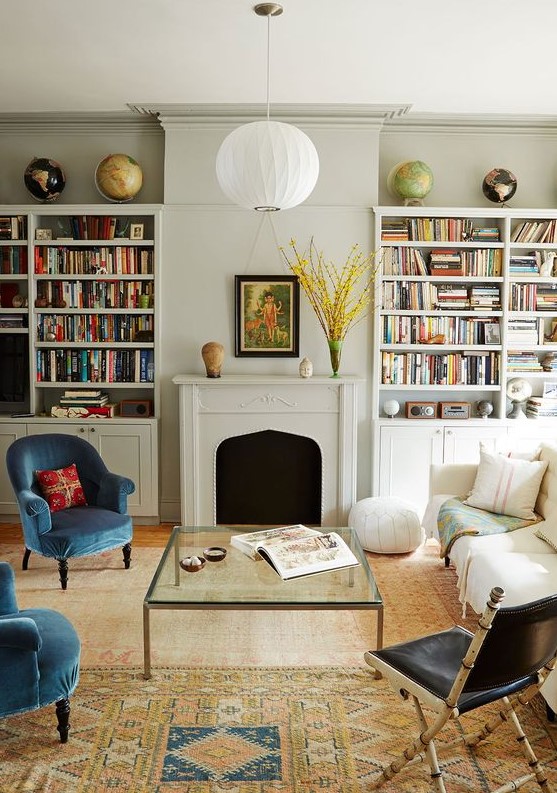 an eclectic living room with built-in bookshelves, a fireplace, a creamy sofa, blue chairs, a black one, a glass coffee table and printed rugs