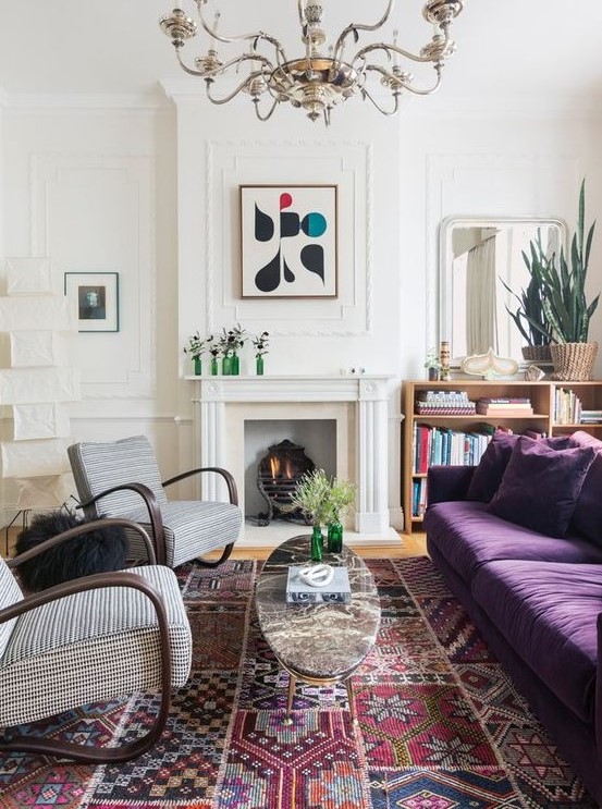 an eclectic living space with a purple sofa, printed rockers, a faux fireplace, a large boho rug and a vintage chandelier