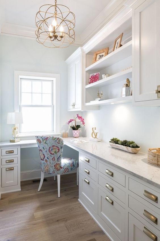 a glam farmhouse home office with white cabinetry, a large L-shaped desk, a printed chair, some decor and a cool gold chandelier