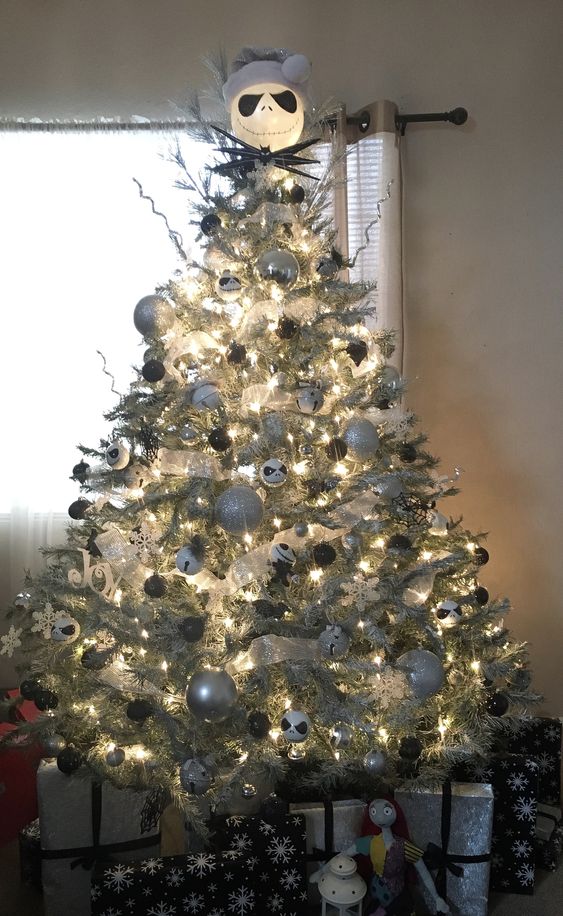 A brightly illuminated silver Christmas tree with silver, black, and white decorations, bells, and a Jack Skellington head on top.