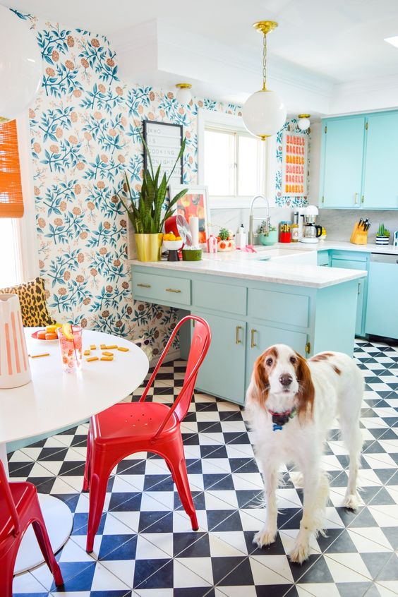 مطبخ كبير ومشرق مع ورق حائط نباتي وخزائن زرقاء فاتحة وطاولة بيضاء وكراسي حمراء بالإضافة إلى أرضية محددة