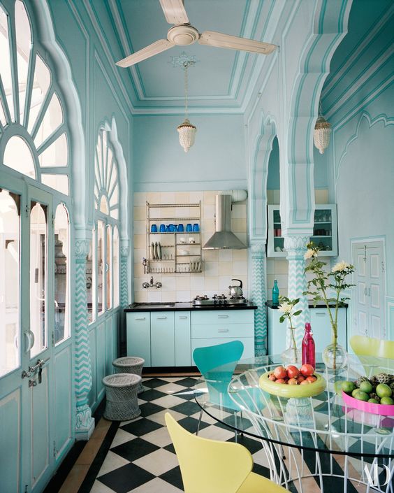 مطبخ على الطراز المغربي باللون الأزرق الفاتح مع أسطح عمل سوداء وأرضية محددة وطاولة زجاجية مستديرة وكراسي صفراء وزرقاء