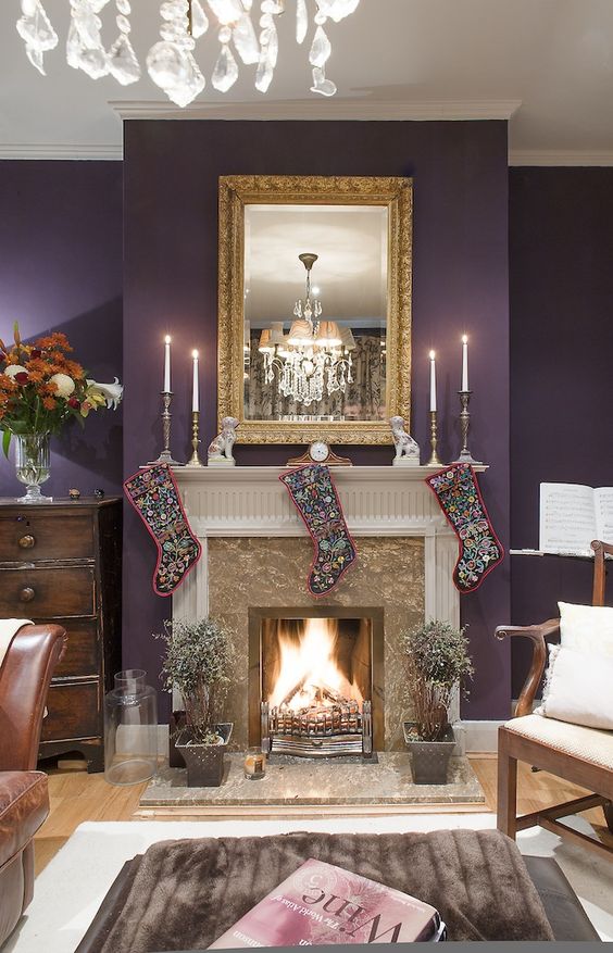 a refined living room with an aubergine accent wall, a fireplace, a mirror in a vintage frame, stockings and a crystal chandelier