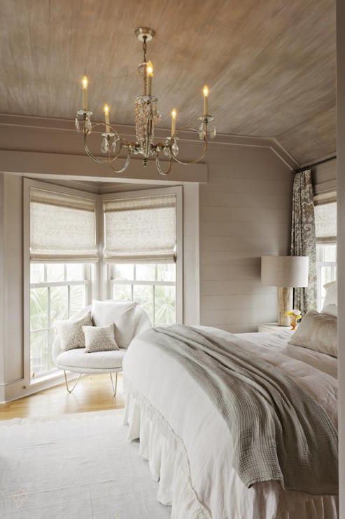 غرفة نوم ريفية مبهجة بجدران مغطاة بألواح خشبية رمادية اللون وسقف وأثاث كريمي وغطاء كريمي وثريا كريستالية راقية