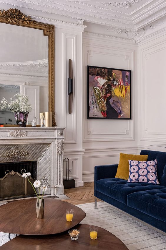 غرفة جلوس باريسية رائعة مع طبقات مزخرفة ومدفأة رخامية راقية ومرآة كبيرة فوقها وأريكة مخملية باللون الأزرق الداكن