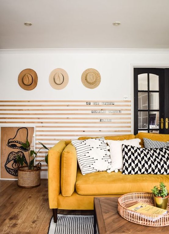 غرفة معيشة أنيقة مع ألواح خشبية على الحائط وأريكة صفراء ووسائد رسومية وقبعات معروضة وبعض النباتات