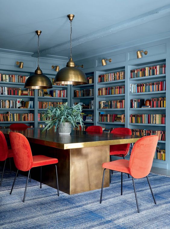 مكتبة منزلية جميلة باللون الأزرق الحجري ومكتب منزلي بها الكثير من الأرفف المدمجة ، ومكتب كبير راقي ، وكراسي حمراء ومصابيح معلقة