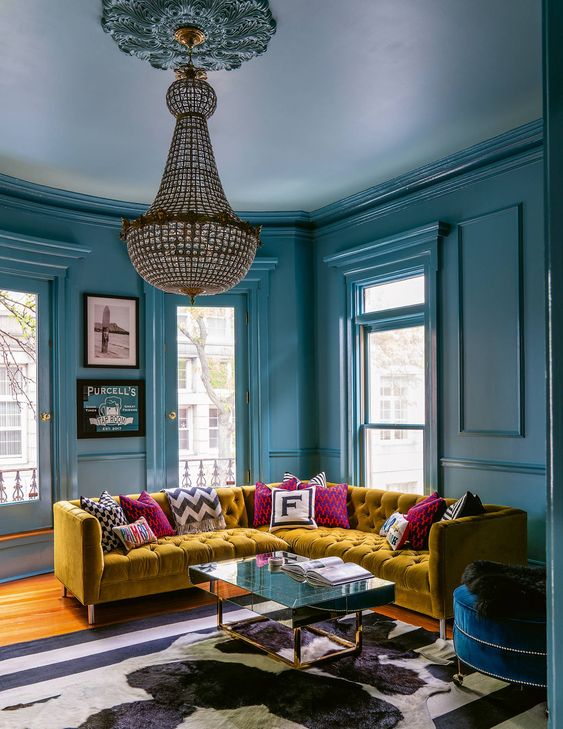 غرفة معيشة مرحة جميلة وجريئة باللون الأزرق مع العديد من النوافذ ، وأريكة زاوية من الخردل ، ووسائد ملونة ومطبوعة ، وثريا من الكريستال