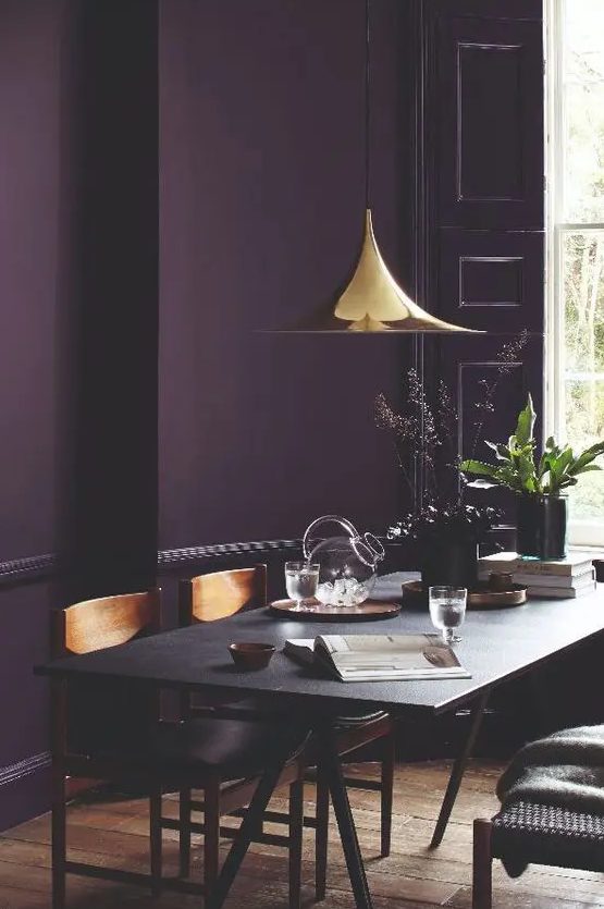 غرفة طعام مزاجية مع جدار بلكنة الباذنجان ، وطاولة سوداء وكراسي خشبية ، ومصباح معلق مذهب لامع