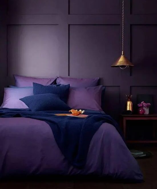 غرفة نوم أحادية اللون مزاجية مع جدار مغطى بألواح أرجوانية عميقة ، وفراش بنفسجي وكحلي ، ومنضدة خشبية ومصباح معلق معدني