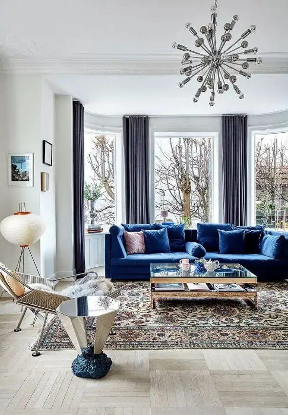 كنب عصري باللون الازرق A-light-filled-living-room-wiht-a-bay-window-a-bold-blue-sofa-a-glass-coffee-table-and-a-mirror-side-table-a-cool-chair-and-a-mid-century-modern-printed-rug