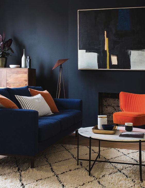 كنب عصري باللون الازرق A-moody-living-room-with-black-walls-a-faux-fireplace-a-navy-loveseat-and-a-bold-orange-chairs-a-round-table-a-statement-artwork-and-some-plants