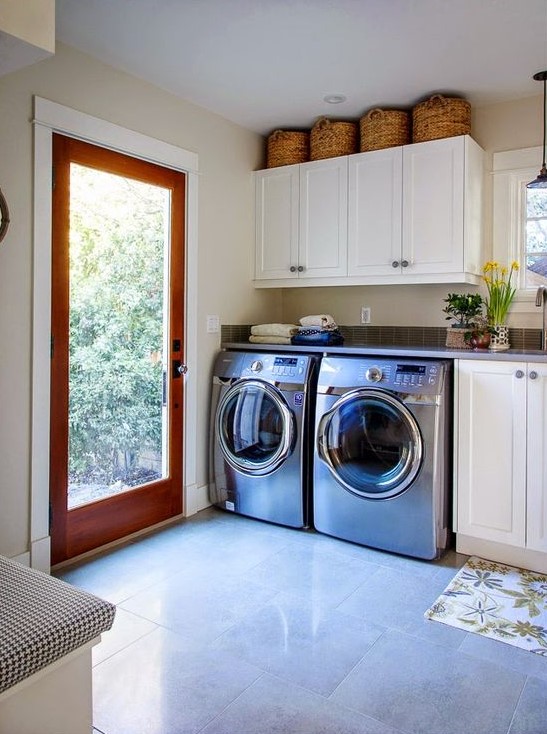 a neutral spacious mudroom laundry with white shaker cabinets, an upholstered bench, a washing machine and a dryer and a glass door