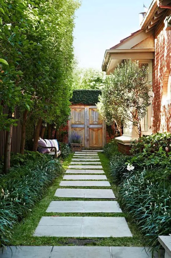 a beautiful and inviting side yard with a green lawn, pavements, greenery and trees, a small bench with pillows and lavender in a basket