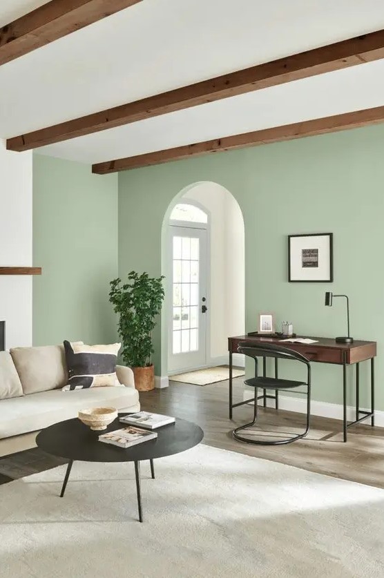 a modern living room with a sage green accent wall, a dark-stained desk and a black chair, a creamy sofa and a black round table