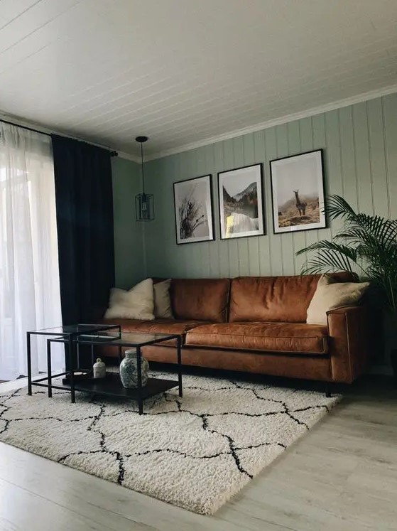 a modern living room with sage green slatted walls, a brown leather sofa, black metal and glass tables and a gallery wall