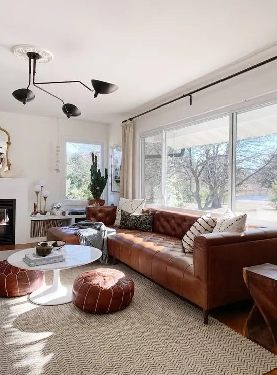 a modern neutral living room with a fireplace, a brown leather Chesterfield sofa, matching ottomans, a black chandelier and a glazed wall