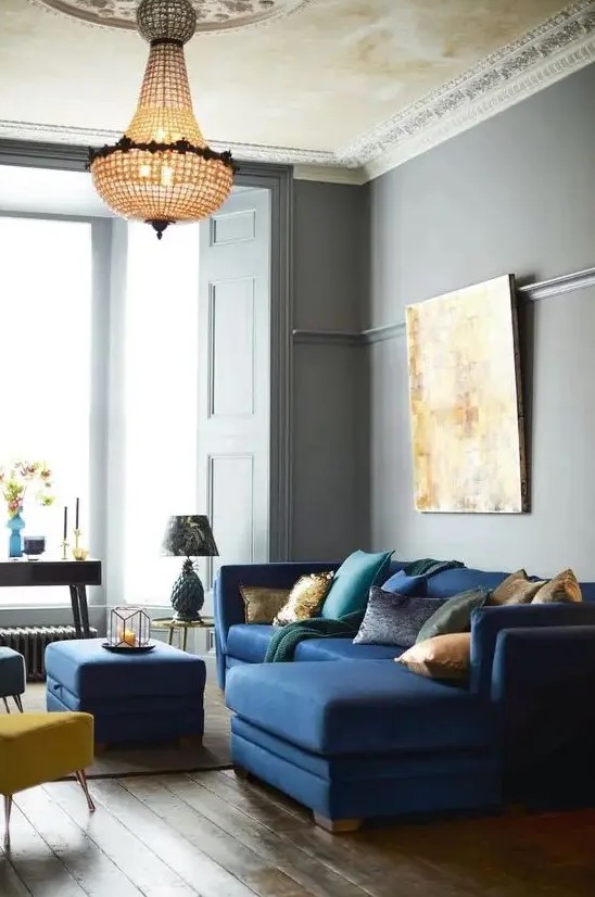 a beautiful and refined living room with grey walls, a navy mid-century modern sectional, a navy pouf and yellow and blue stools, a crystal chandelier and a statement artwork