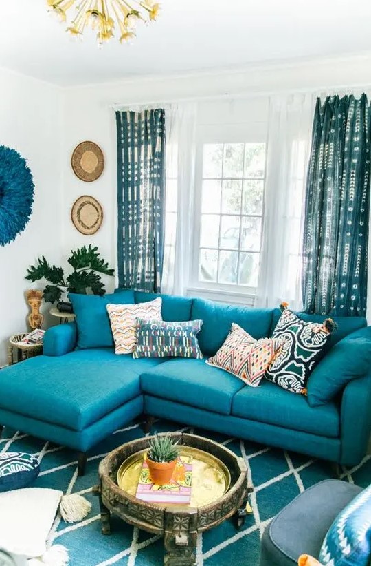 a bright living room with a turquoise sofa, a turquoise printed rug and curtains, decor on the wall and a carved wooden coffee table